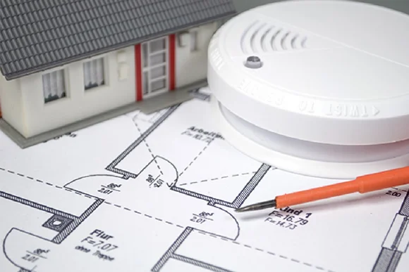 Ein Rauchmelder liegt auf einem Bauplan neben einem Modellhaus und einem Schraubenzieher.